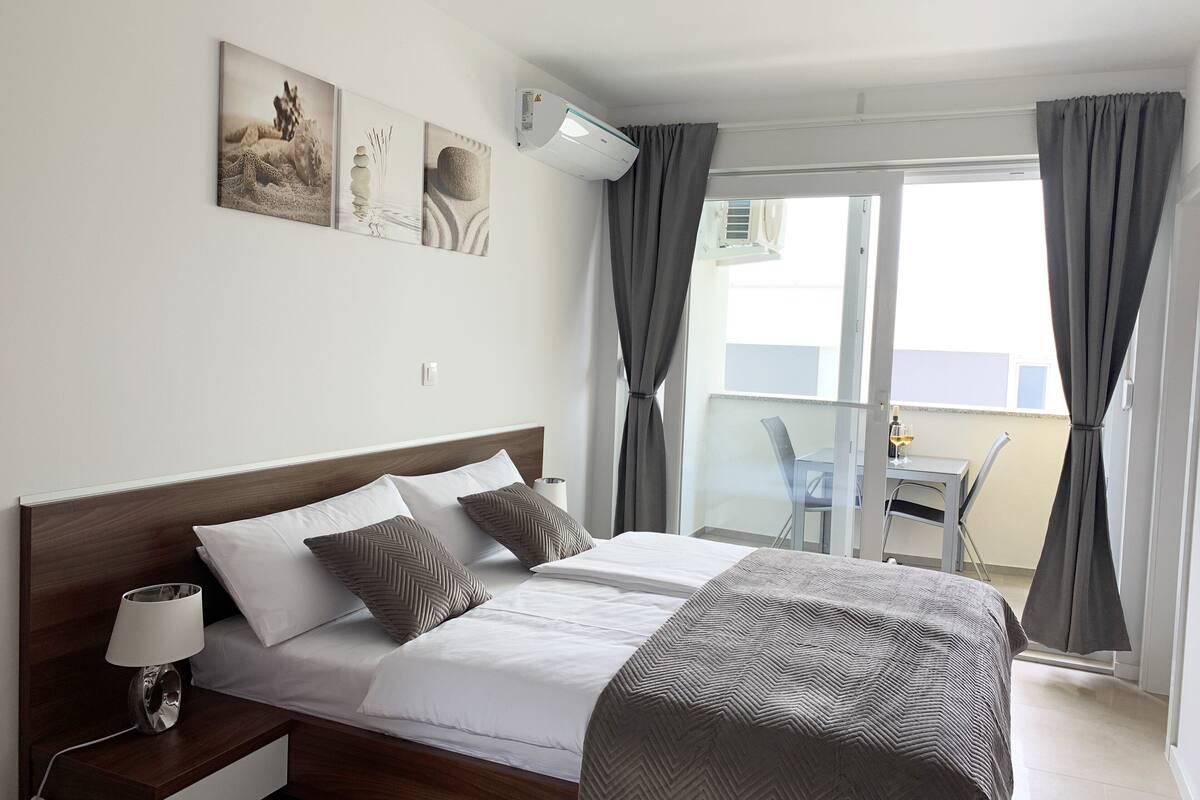 A hotel room with a bed, bedside table, lamp, air conditioning unit, and artwork on the wall.