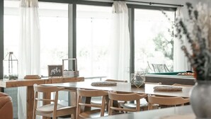Een eetgedeelte met een houten tafel en stoelen, grote ramen met dunne gordijnen en uitzicht op groen.