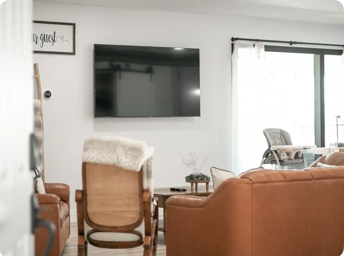 Ein Wohnzimmer mit Flachbildfernseher, einem braunen Ledersofa, einem Holzstuhl mit weißer Decke und einem Essbereich mit Tisch und Stühlen.