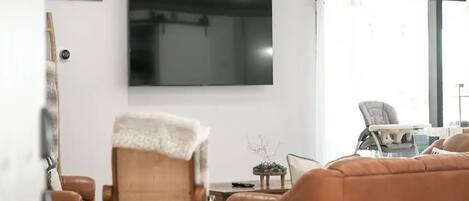 Ein Wohnzimmer mit Flachbildfernseher, einem braunen Ledersofa, einem Holzstuhl mit weißer Decke und einem Essbereich mit Tisch und Stühlen.