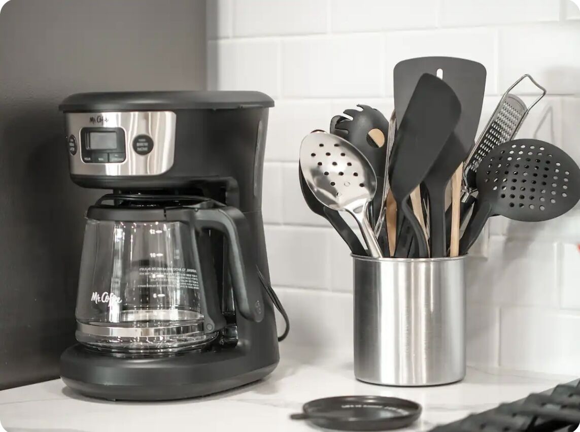 Una cafetera con pantalla digital, un vaso de acero inoxidable y utensilios, y una encimera de cocina.