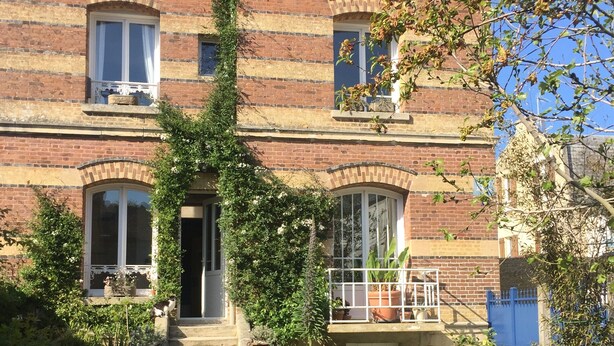 Una casa a due piani in mattoni con un ingresso principale, un piccolo portico e un giardino con piante in vaso.