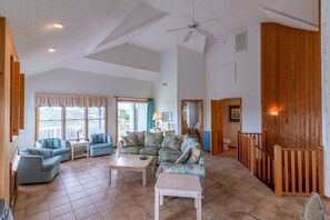 A spacious living room with a large sofa set, a coffee table, and a television. There is a wooden staircase and a wooden wall paneling on one side. - Cozy cottage between the ocean and the sound in Duck, NC by Sun Realty (Duck)