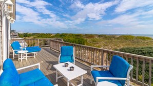 Eine Holzterrasse mit blauen Sitzgelegenheiten und einem kleinen Tisch, mit Blick auf einen Strand und Dünen.