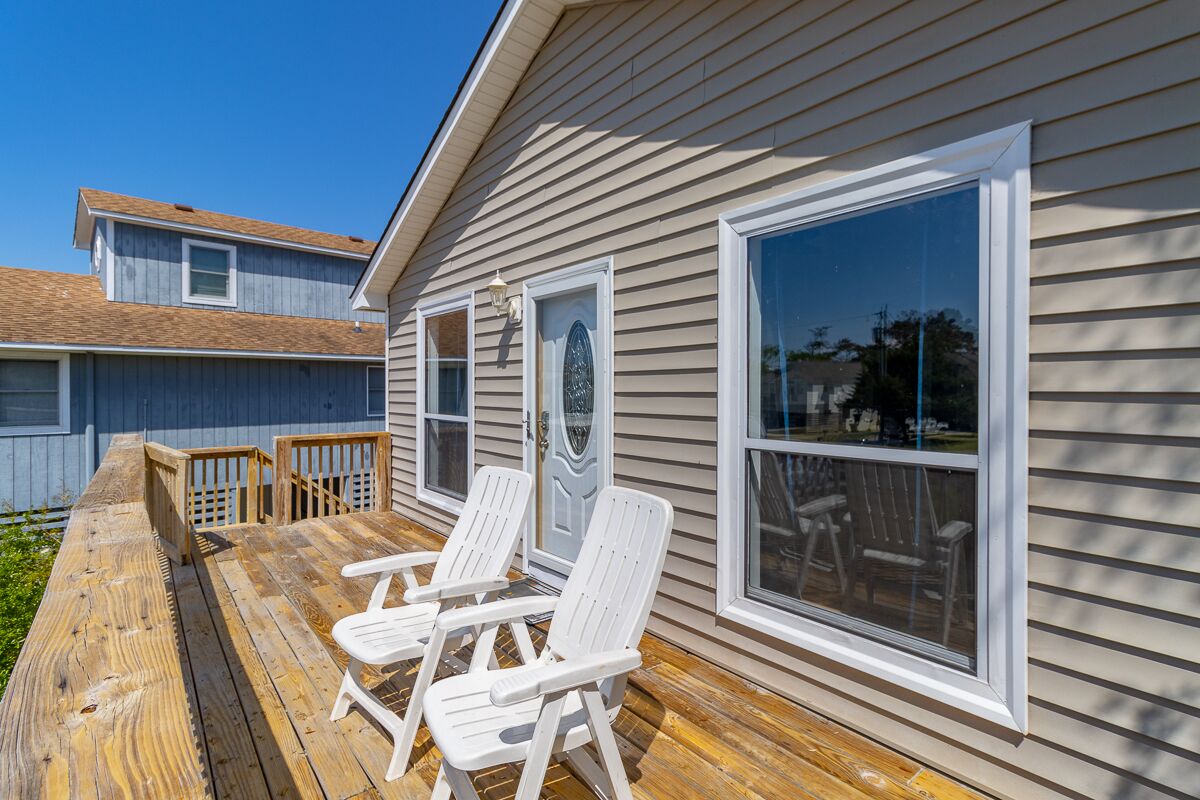 A wooden deck with two white Adirondack chairs, a wooden railing, and a house with large windows and a door with a decorative glass panel.