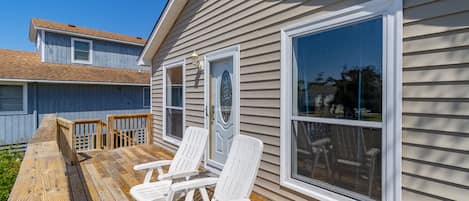 Una terraza de madera con dos sillas Adirondack blancas, una barandilla de madera y una casa con ventanas grandes y una puerta con un panel de vidrio decorativo.
