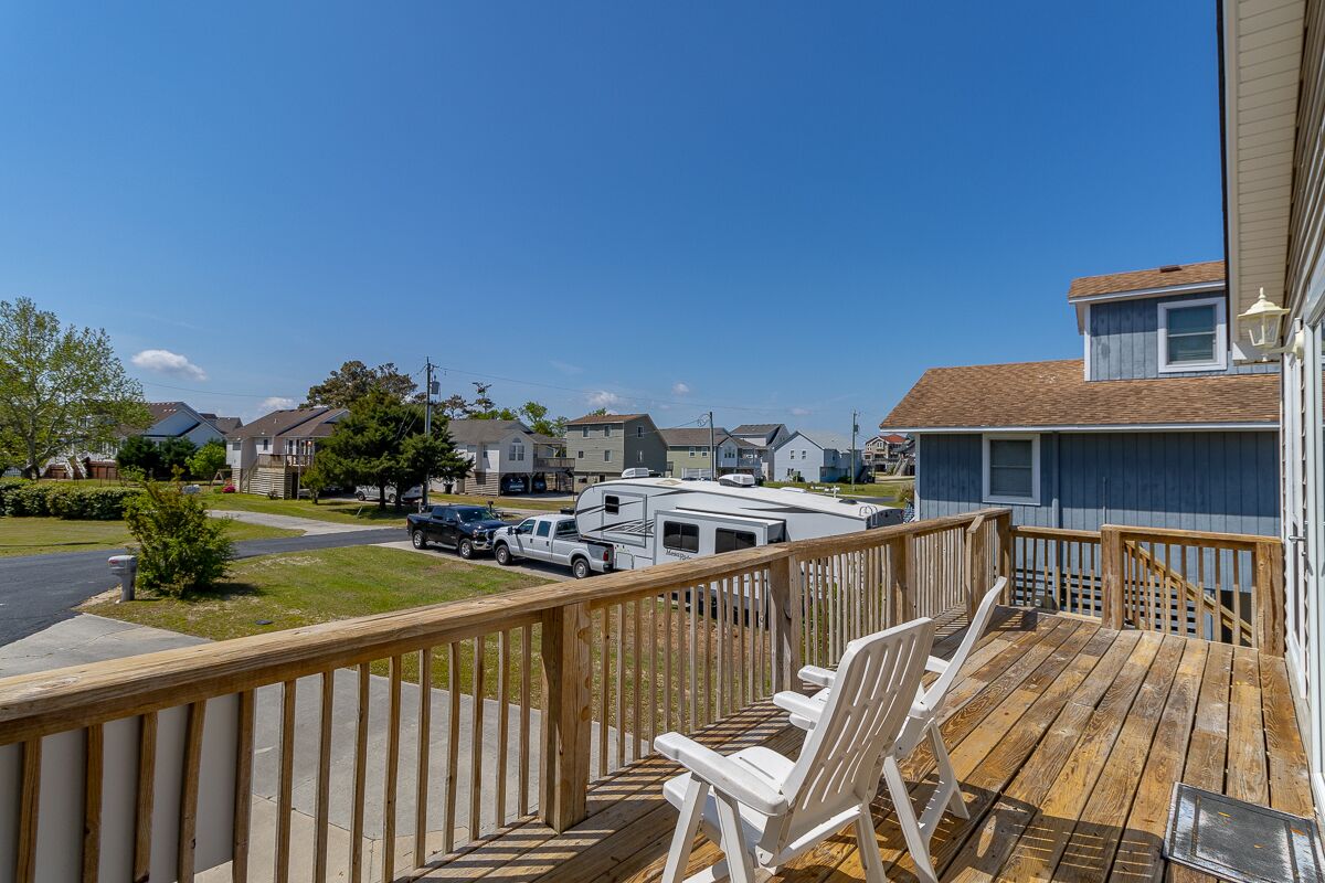 A wooden deck with white chairs, a clear view of a residential street, and a large RV parked in the background.