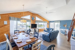 A spacious living area with a dining table set for four, a blue sofa, and a television on a stand. - Sunrise views, volleyball court and a quick walk to the beaches by Sun Realty (Duck)
