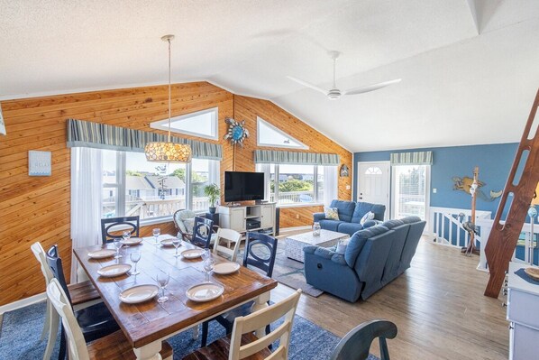 A dining area with a wooden table and chairs, a blue sofa, and a TV in a room with wooden walls and large windows.