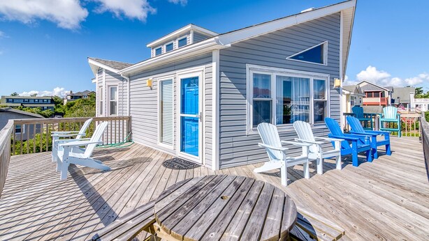 Een huis aan zee met een terras, met een klein gebouw met een blauwe deur en ramen, omringd door witte en blauwe Adirondack stoelen.