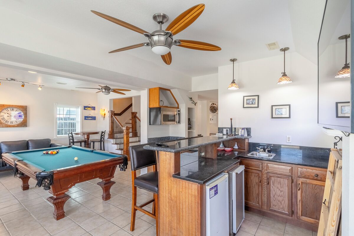 A spacious living area with a pool table, a ceiling fan, and a kitchen area with wooden cabinets and a refrigerator.
