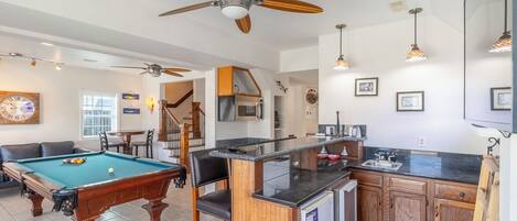 A spacious living area with a pool table, a ceiling fan, and a kitchen area with wooden cabinets and a refrigerator.