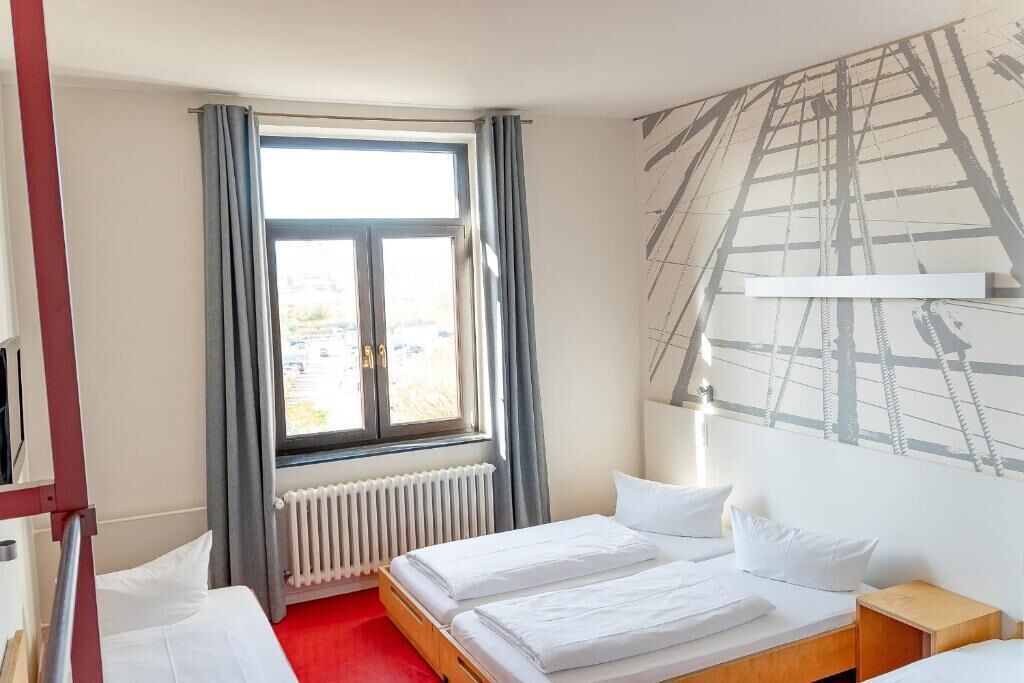 Habitación de hotel con dos camas, una ventana con cortinas, un radiador y una pared con un mural de temática náutica.