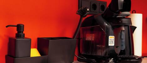 A room with a red wall, a black vacuum cleaner, a black coffee maker, a black hairdryer, and a black hairbrush.