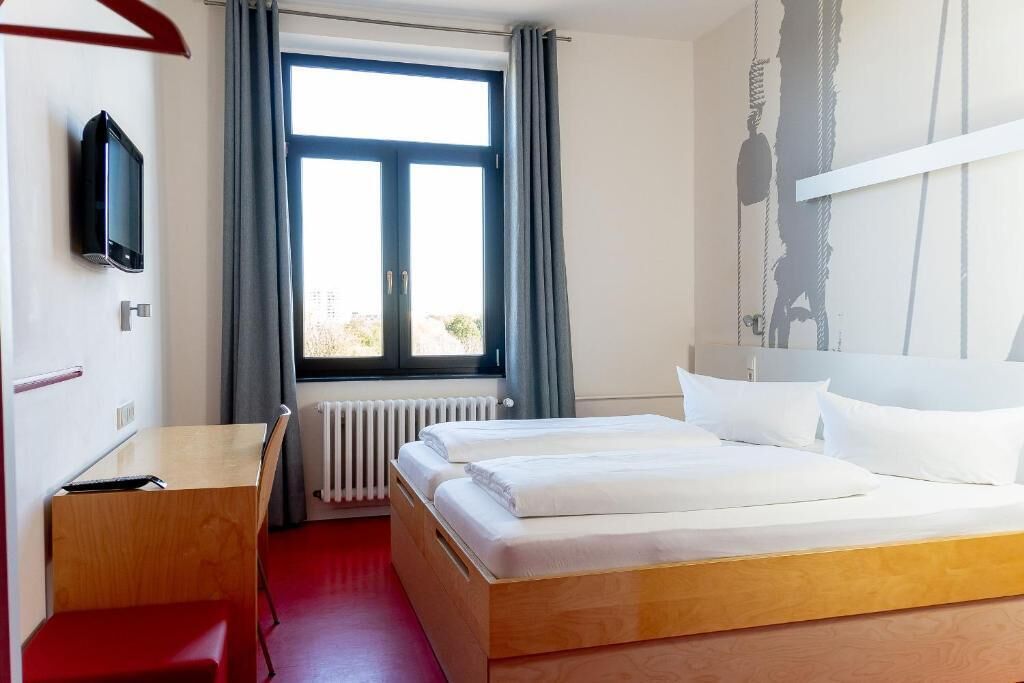 A hotel room with a bed, a desk, a chair, a TV, a window with curtains, and a radiator.