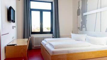 A hotel room with a bed, a desk, a chair, a TV, a window with curtains, and a radiator.