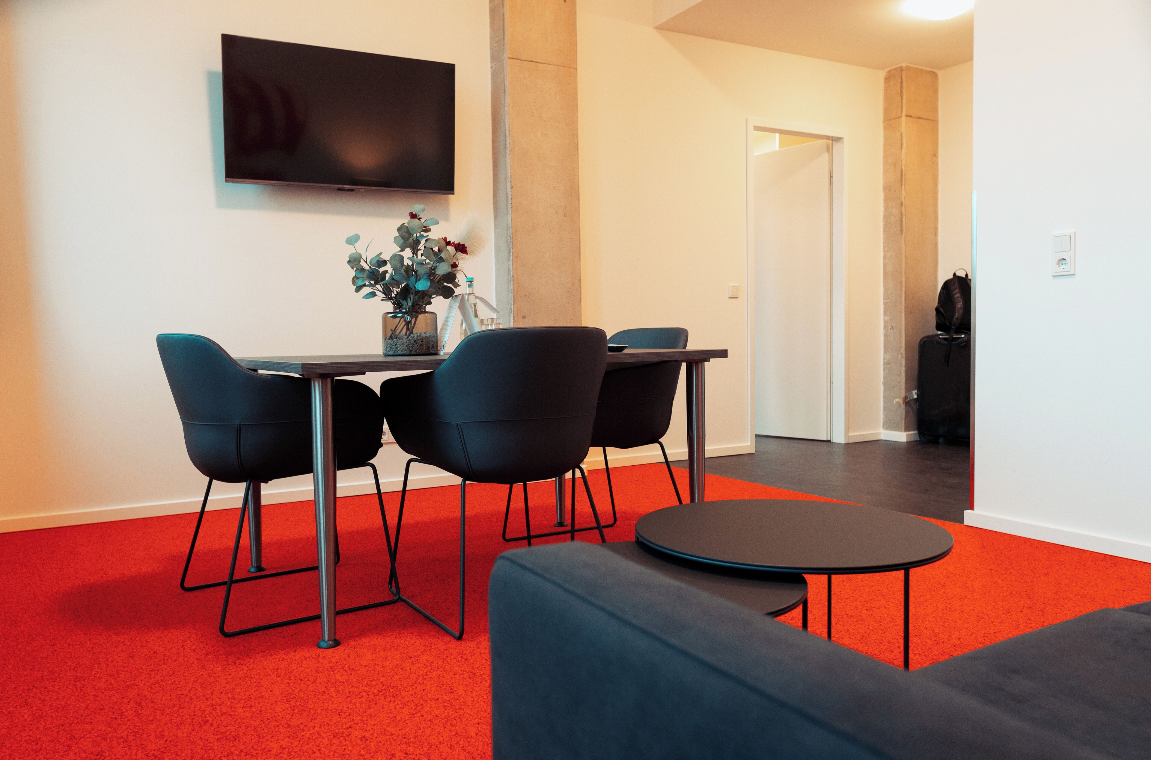 Een moderne woonkamer met een rode vloerkleed, een flatscreen televisie aan de wand, een eettafel met vier stoelen en een bank.