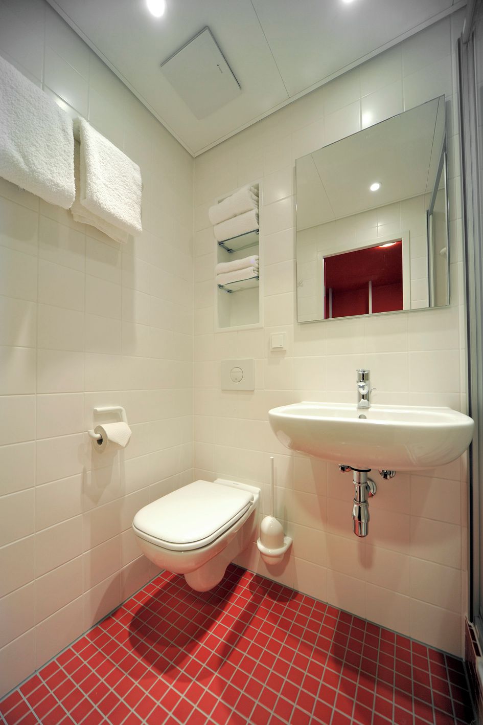 A small bathroom with a toilet, sink, and mirror. There are white towels hanging on the wall, and a red-tiled floor.