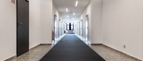 Un pasillo largo y estrecho con alfombra negra, paredes blancas y una puerta con el número 319 a la izquierda.