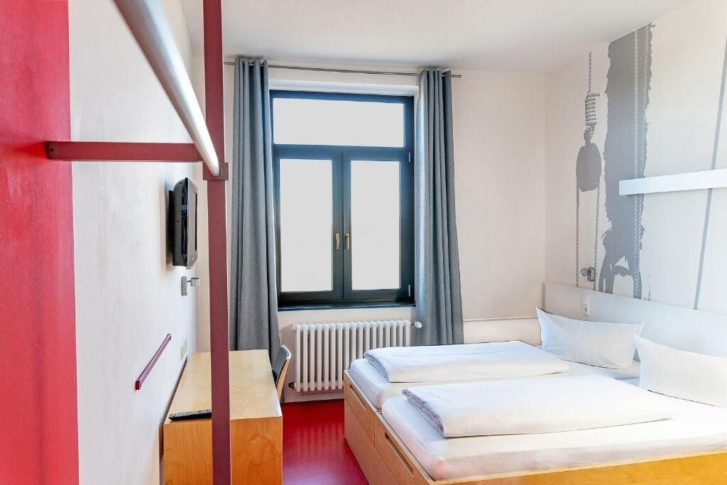 Habitación de hotel con cama, escritorio, radiador, ventana con cortinas y televisor montado en la pared.