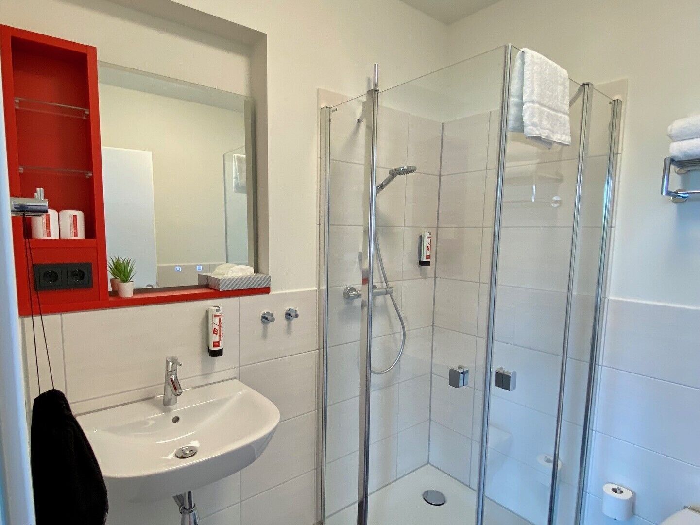 Une salle de bain moderne avec une douche à parois de verre, un lavabo blanc et une étagère rouge où sont rangés les articles de toilette.