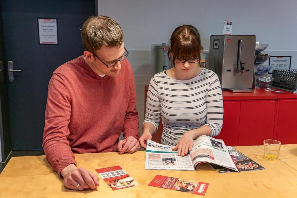 Deux personnes sont assises à une table, devant un magazine et un verre, avec un réfrigérateur en arrière-plan.