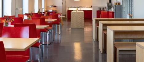 A modern dining area with red chairs and tables, a central island, and a kitchen in the background.