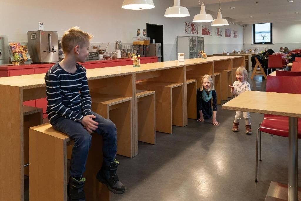 A modern cafeteria with wooden tables and benches, a refrigerator, and a coffee station.