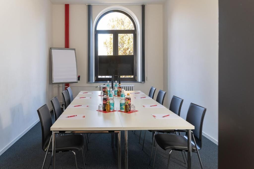 Ein Konferenzraum mit langem Tisch, schwarzen Stühlen, einer Tafel und einem Fenster mit Blick auf Bäume.