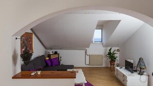 Un moderno loft con soffitto a volta, una zona salotto accogliente con divano e cuscini, un piccolo tavolo da pranzo, una TV e un termosifone.