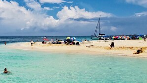 Ein Strand mit klarem türkisblauem Wasser, darin schwimmende Menschen und bunten Sonnenschirmen im Sand.