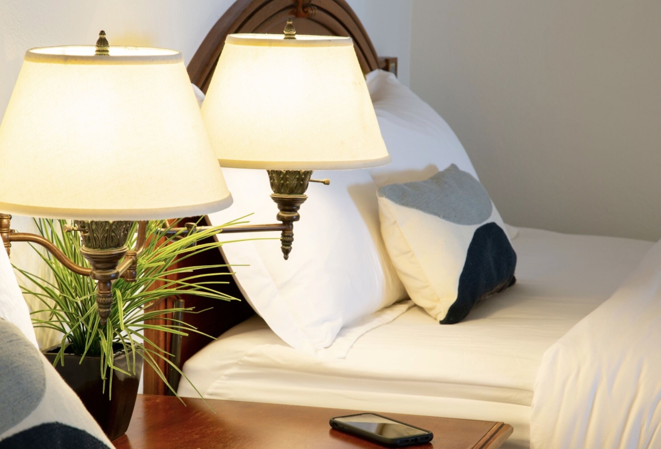 Un lit bien fait, avec deux lampes de chevet, un téléphone intelligent posé sur la table de nuit et une plante en pot.