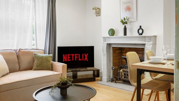 A living room with a beige sofa, a coffee table with a plant, a TV displaying 'NETFLIX', a fireplace, and a dining area with chairs and a table. - 2 single or 1 King bed in Notting Hill.. Super location for sightseeing London
 (London)
