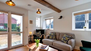 Un salon chaleureux avec un canapé, des fauteuils et une table basse. De grandes fenêtres sont équipées de stores, une porte donne sur l’extérieur et le plafond est orné de poutres apparentes.
