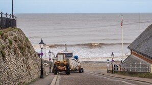 A coastal road with a vehicle parked near the sea, a stone wall, and a building with a sloped roof. - The Old Sunday School & Vestry (Cromer)