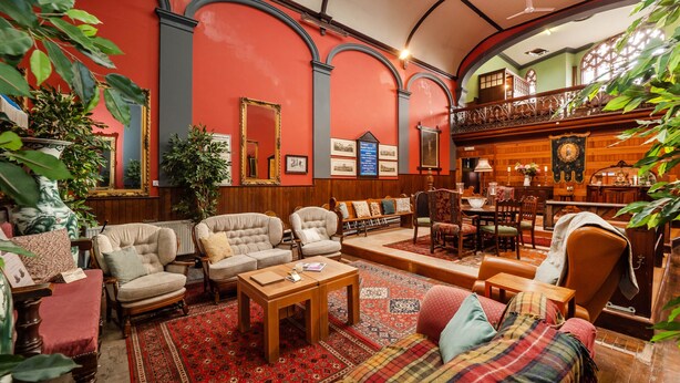 A spacious lobby with a high ceiling, red walls, and wooden paneling. There are multiple seating areas with sofas and armchairs, a coffee table, and a dining table. The lobby features a large mirror, framed pictures, and potted plants. - The Old Sunday School & Vestry (Cromer)