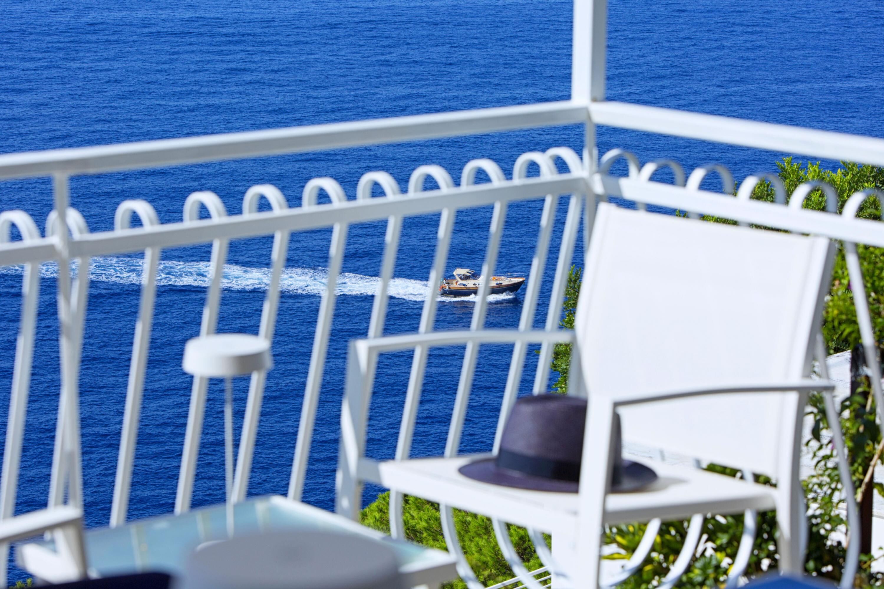 A balcony with a white railing, a table, and a chair. A boat is visible on the water in the background.