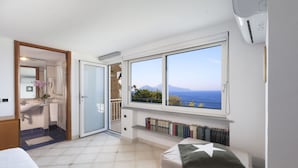 A bedroom with a large window, a bed, a bedside table, a bookshelf, and a bathroom with a shower.