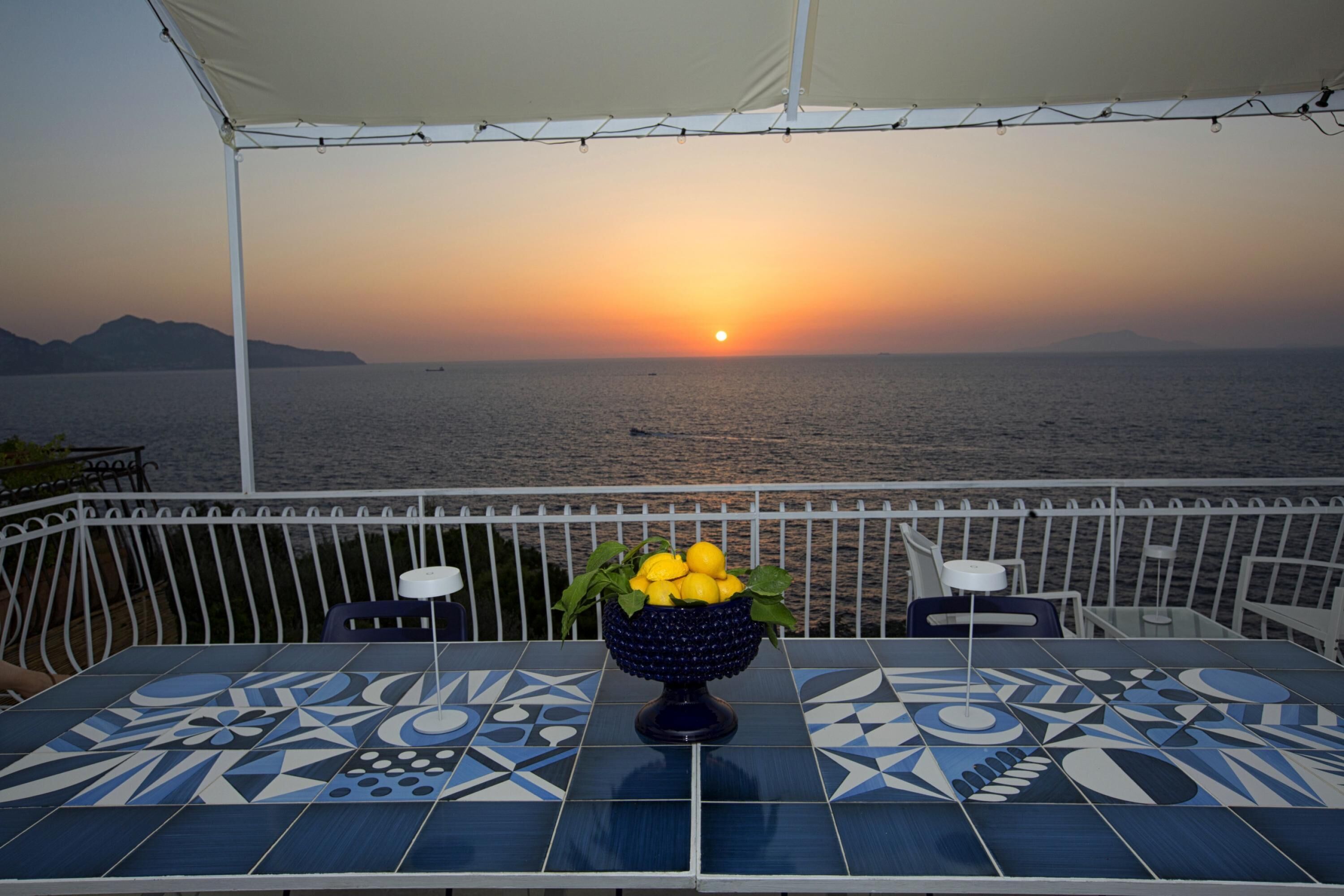 A table with a decorative bowl of lemons and mint leaves, set against a backdrop of a sunset over the sea, with a tiled floor and white railing.