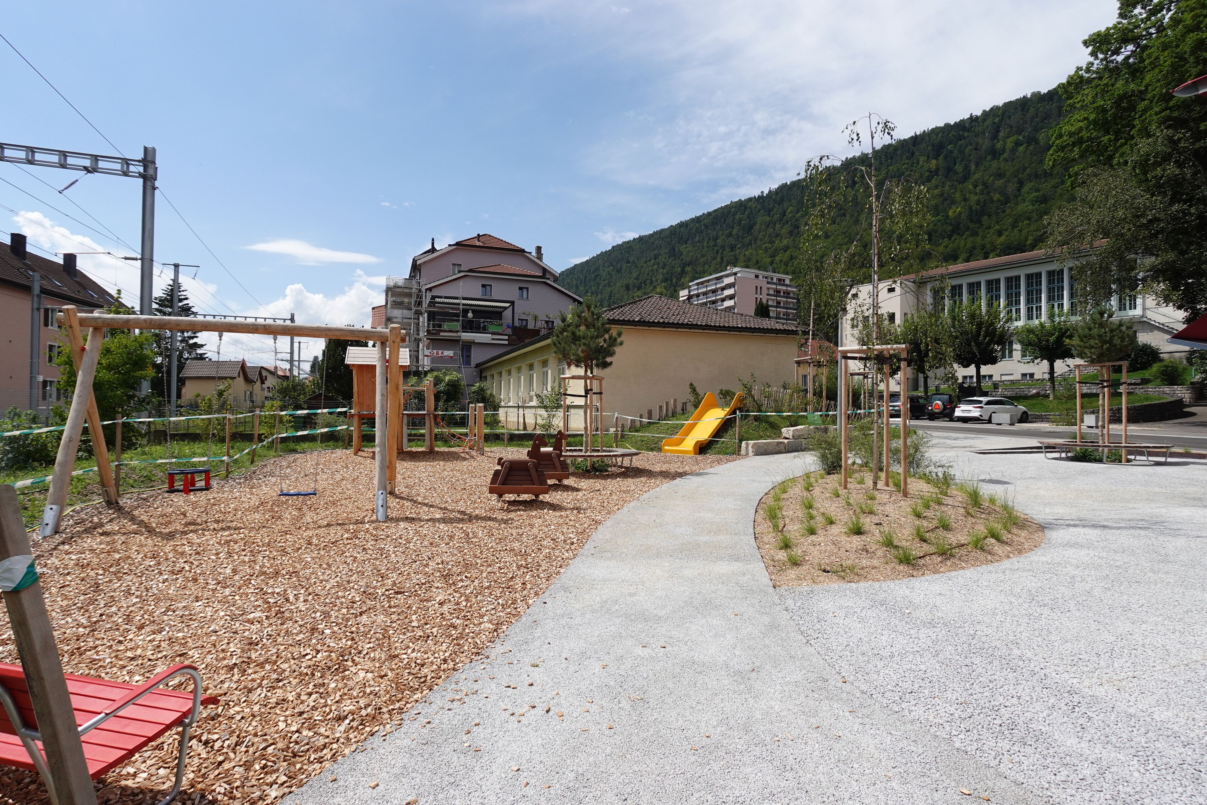 Un parque infantil con tobogán y columpios, rodeado por un sendero de grava y una pequeña zona ajardinada.