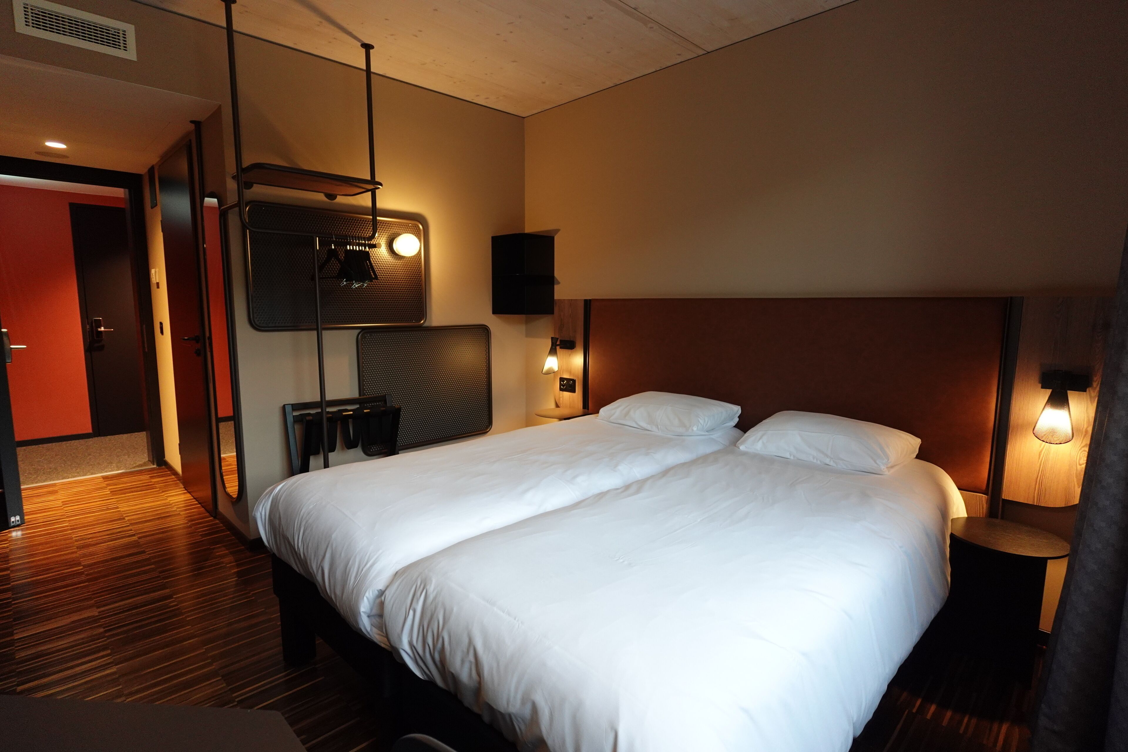 A hotel room with a large bed, a wooden floor, a wall-mounted TV, and a bedside table with a lamp.
