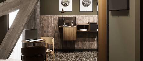A modern hotel room with a sloped ceiling, a bed, a desk with a laptop, a chair, a small kitchen area, and artwork on the wall.