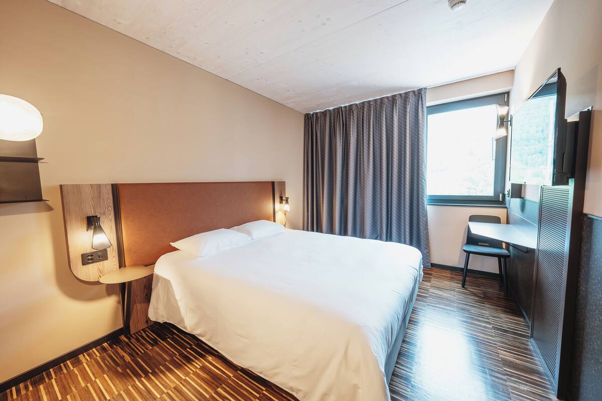 Une chambre d’hôtel avec un grand lit, un bureau, une chaise, un miroir et une fenêtre avec des rideaux.