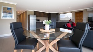 A modern kitchen with dark cabinets, a stainless steel refrigerator, and a dining area with a glass table and black chairs. - Ocean View Executive Apartment 2 (Geographe)