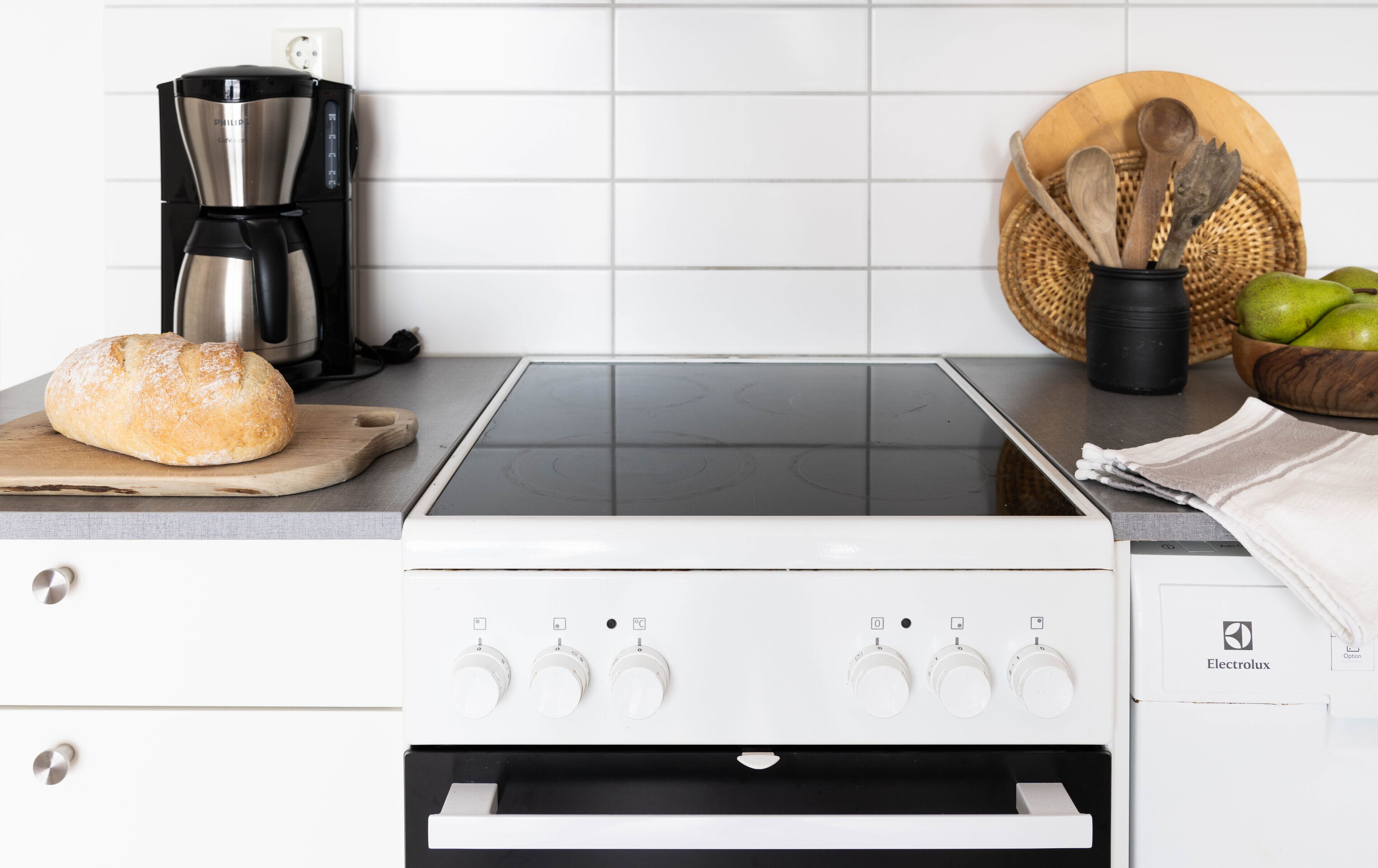 Una cocina con encimera blanca, un pan, una cafetera y una cesta de mimbre con utensilios de madera.