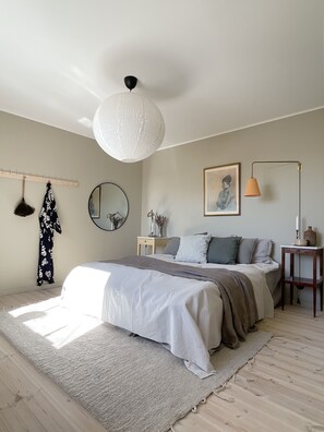 A bedroom with a large bed, a hanging lamp, a bedside table, a mirror, and a framed picture on the wall. - Hönö Sjöbodar (Hono)
