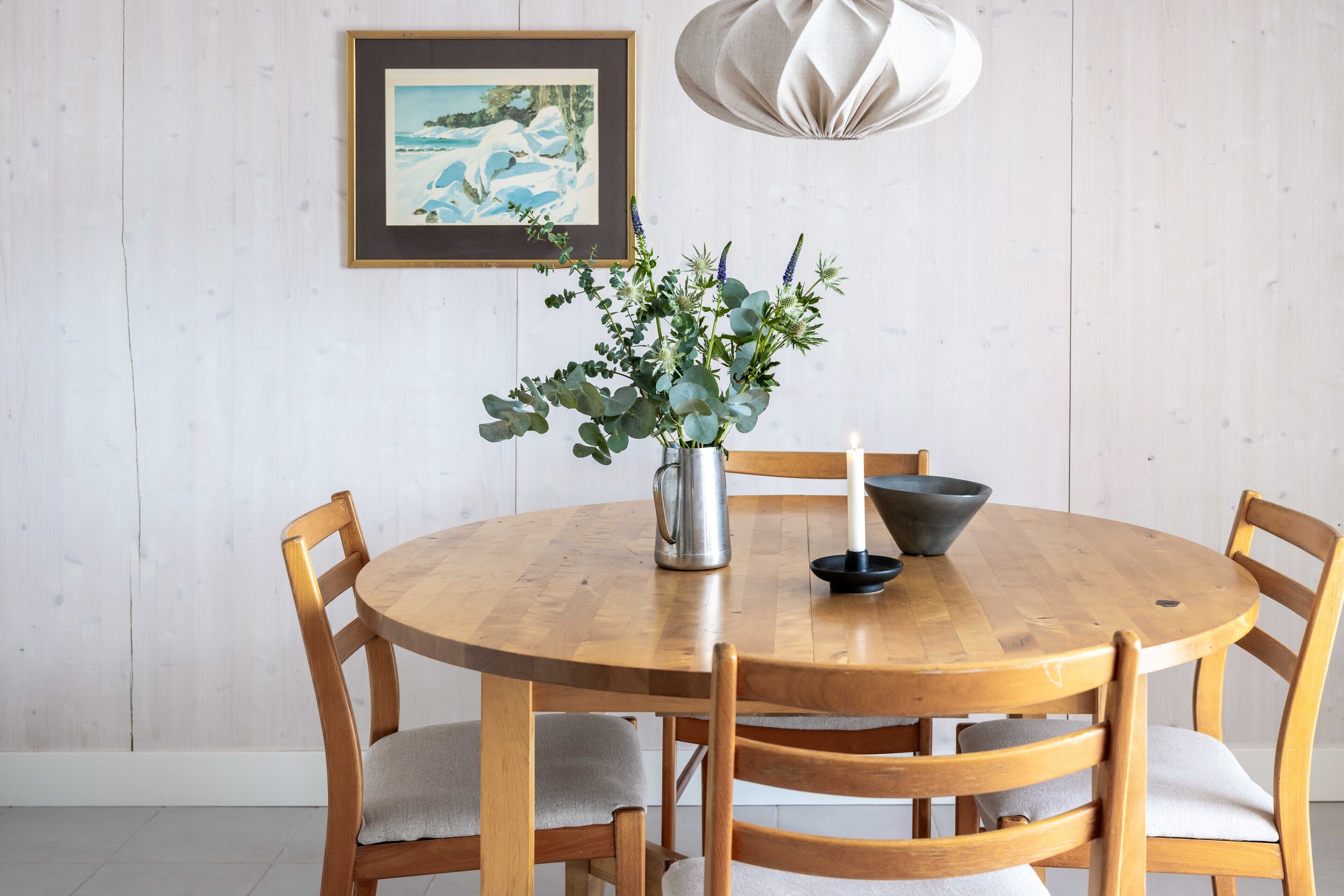 A wooden dining table with six chairs, a vase with greenery, a candle, and a framed painting on the wall.