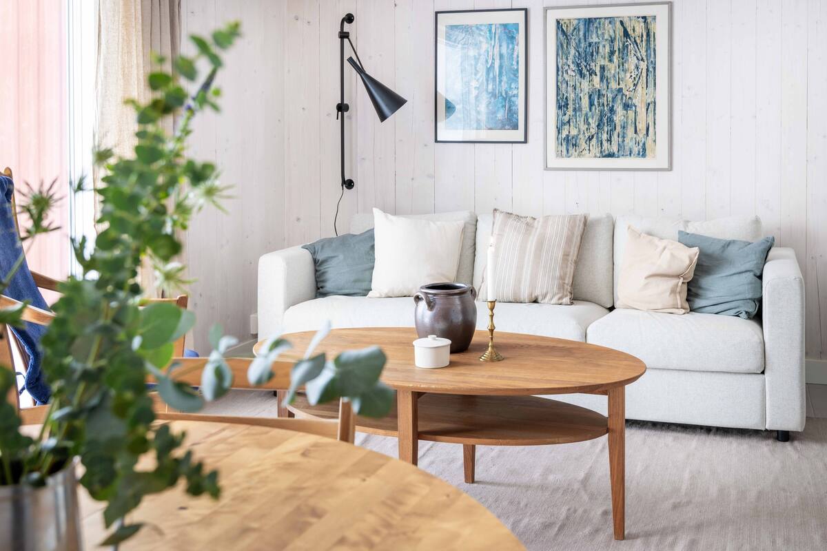 A living room with a white sofa, a wooden coffee table, and a vase with candles. There are two framed artworks on the wall and a plant in the foreground.