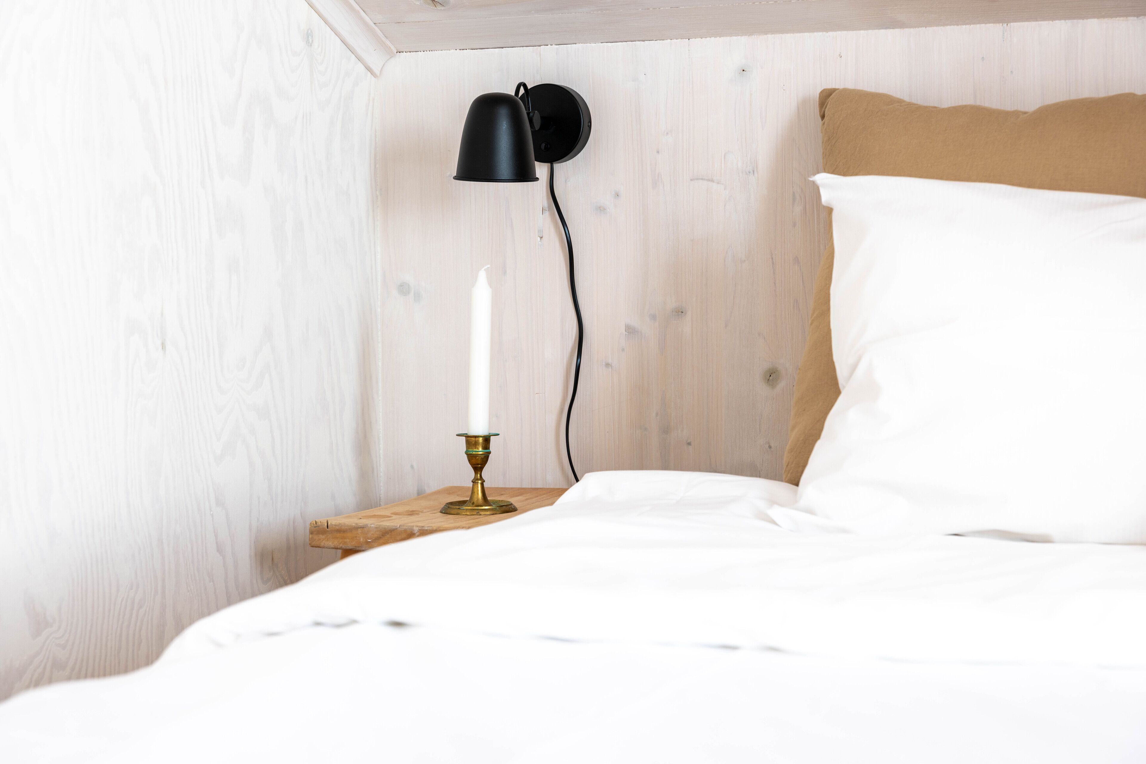 Een net opgemaakt bed met witte lakens, een houten nachtkastje, een messing kandelaar met één witte kaars, en een zwart wandlamp.