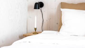 Een net opgemaakt bed met witte lakens, een houten nachtkastje, een messing kandelaar met één witte kaars, en een zwart wandlamp.